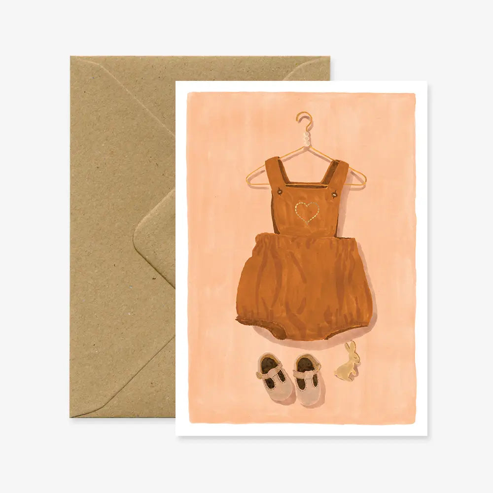 Brauner Baby-Latzhose mit Herzmuster auf einem Kleiderbügel, dazu kleine rosa Schuhe und ein Hasenanhänger auf beigem Hintergrund