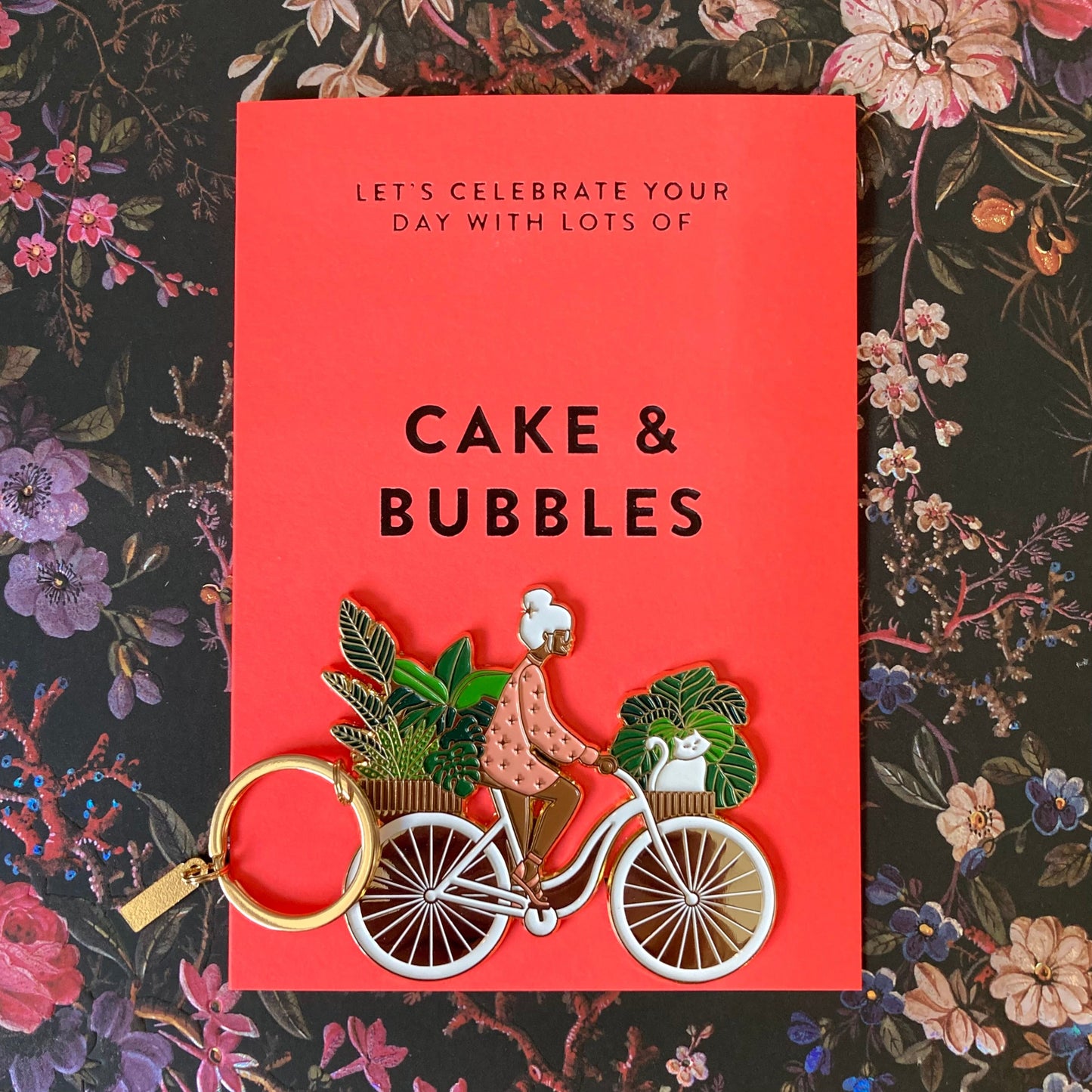 Rote Karte mit dem Text 'Let's celebrate your day with lots of cake & bubbles' und einem Schlüsselanhänger in Form einer Person auf einem Fahrrad mit Pflanzen und einer Katze