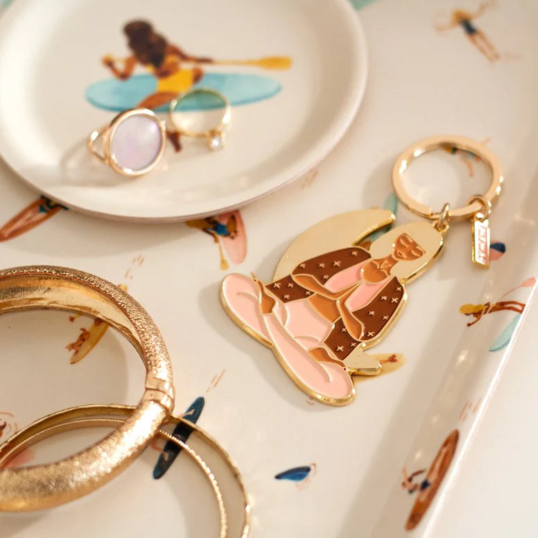 Goldfarbener Schlüsselanhänger mit einer sitzenden Frau in rosa Kleidung auf einem Tablett und goldfarbenem Schmuck