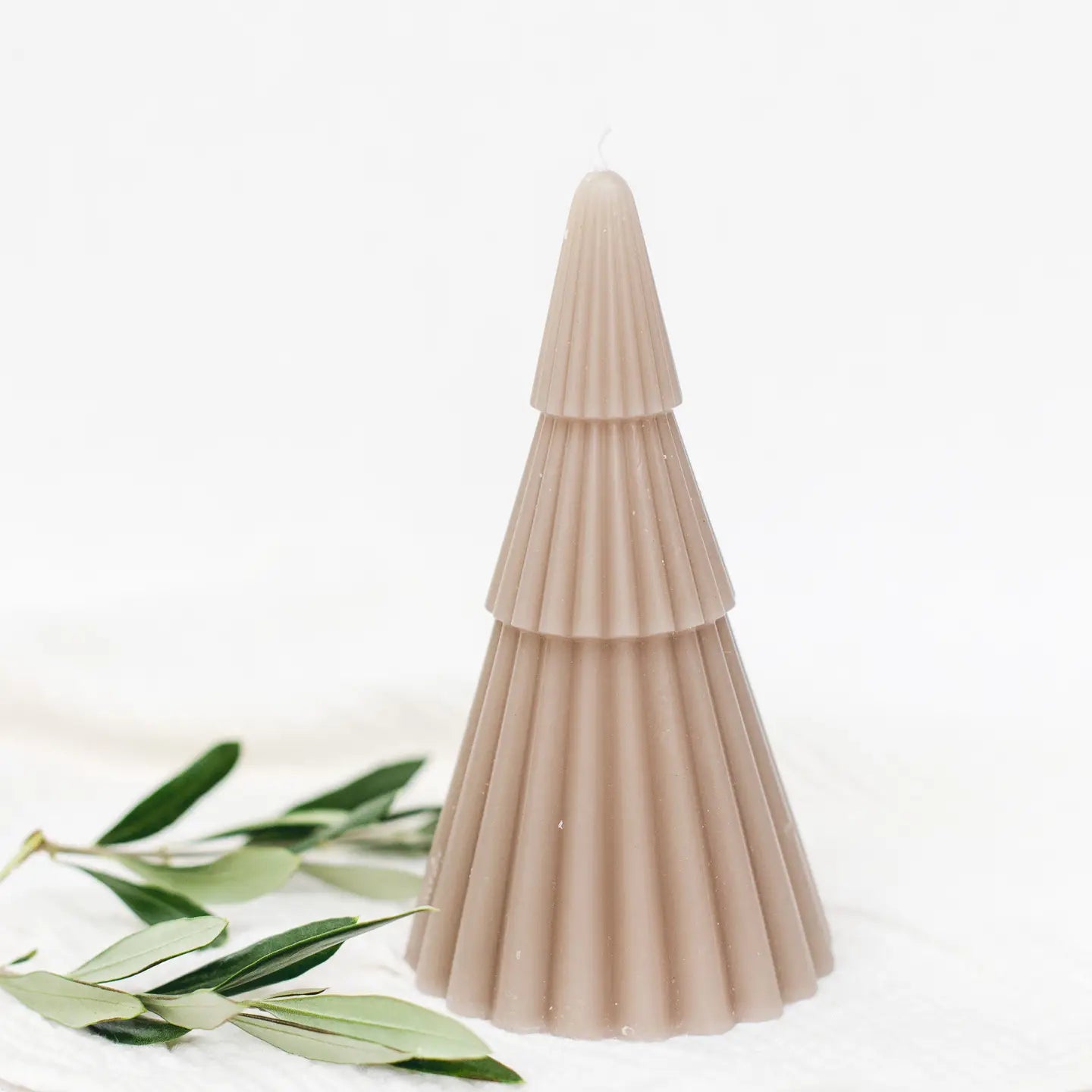 Beige Kerze in Form eines gestuften Tannenbaums mit gerillter Oberfläche, daneben grüne Blätter.