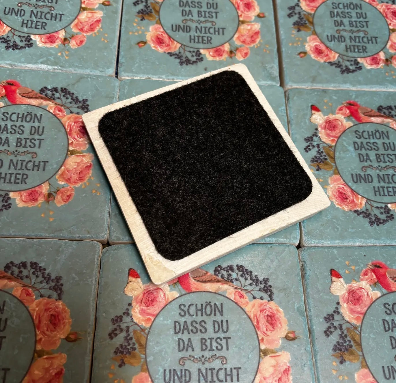 imVordergrund fliese von hinten mit filz, Türkisfarbene Untersetzer mit rosa Rosen, Vogel und Schmetterling, Text 'Schön dass du da bist und nicht hier'