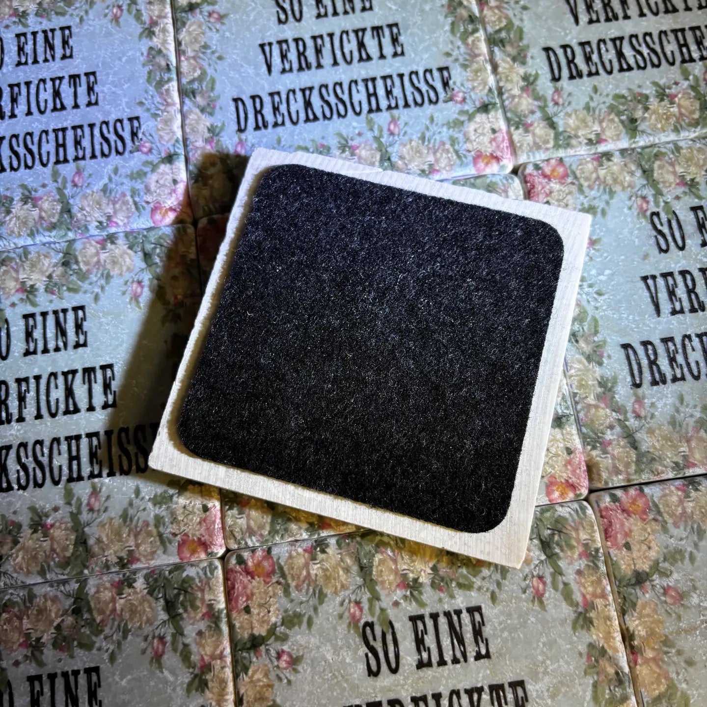 Quadratische Fliese von hinten mit filz, im Hintergrund fliesen mit Blumenmuster und dem Text 'SO EINE VERFICKTE DRECKSSCHEISSE' in Großbuchstaben