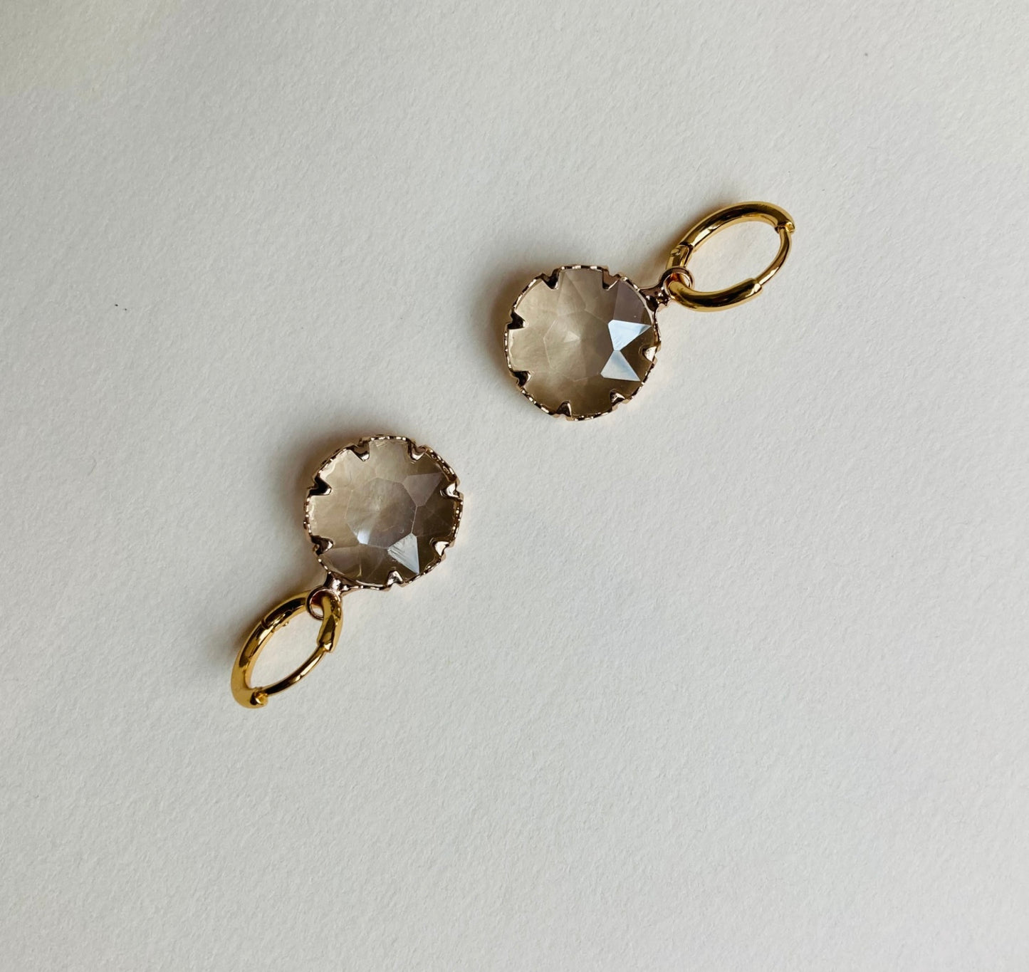 Ohrringe mit goldfarbenen Creolen und facettierten, runden, transparenten Anhängern auf hellem Untergrund