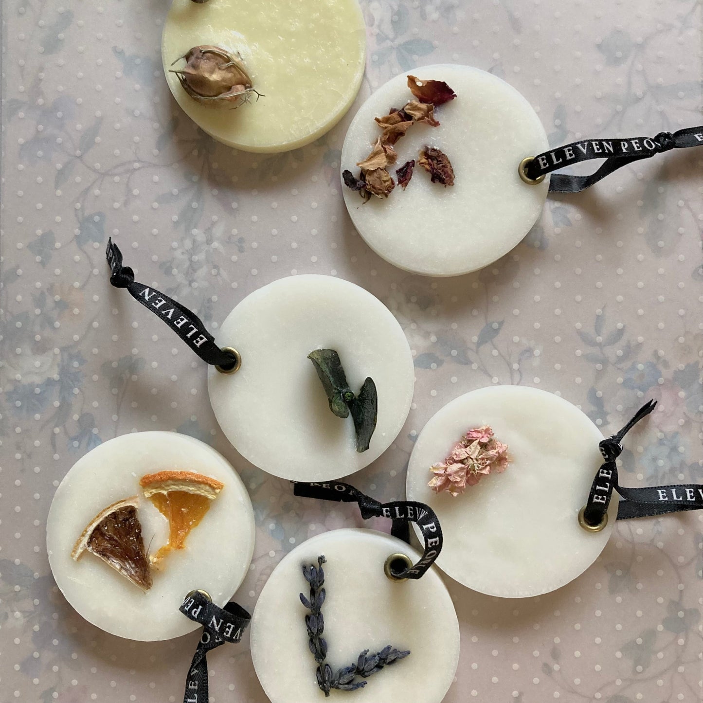 runde, weiße Sojawachs-Lufterfrischer von ELEVEN PEOPLE auf beigem, gepunktetem Hintergrund. Jeder Lufterfrischer zeigt verschiedene getrocknete Pflanzenelemente, die in die Wachsoberfläche eingebettet sind Sie haben schwarze Bänder mit dem Aufdruck „ELEVEN PEOPLE“.