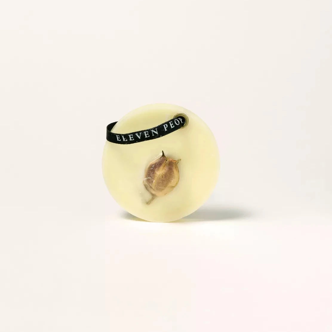 Ein runder, weißer Sojawachs-Lufterfrischer von ELEVEN PEOPLE auf beigem, gepunktetem Hintergrund. Der Lufterfrischer zeigt ein getrocknetes Pflanzenelement, dass in die Wachsoberfläche eingebettet ist. Er hat ein schwarzes Band mit dem Aufdruck „ELEVEN PEOPLE“