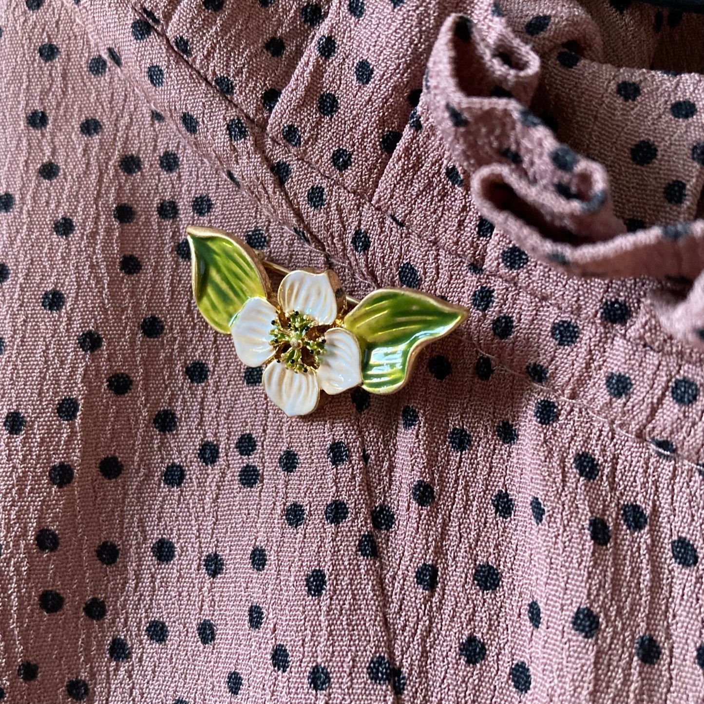 Goldfarbene Brosche in Form einer Blüte mit weißen Blütenblättern und zwei grünen Blättern, verziert mit kleinen grünen Punkten in der Mitte an einem gepunkteten Stoff.