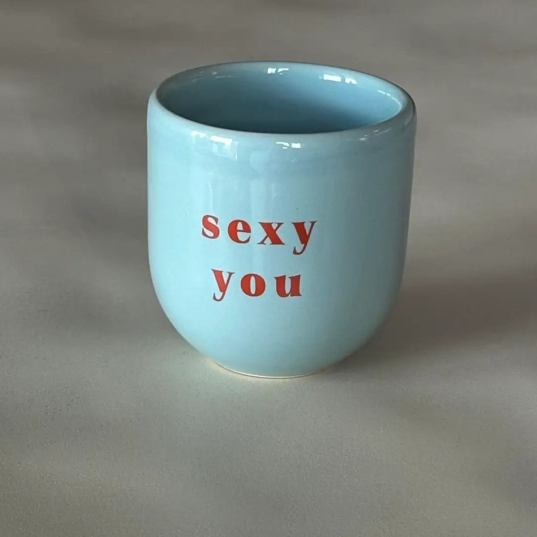 Ein hellblauer Keramikbecher mit weißer Aufschrift „Sexy you“ auf hellem Hintergrund.