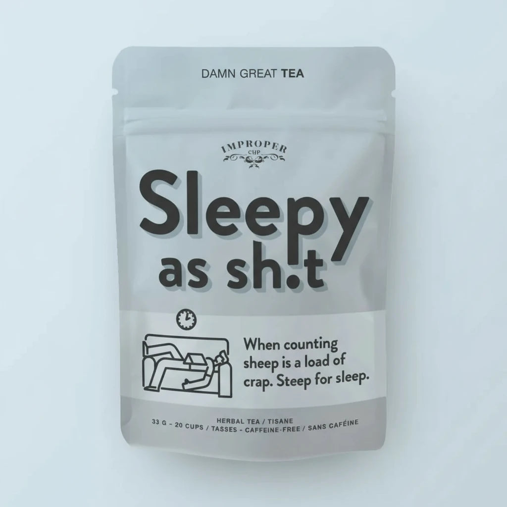 Graue Verpackung mit schwarzer Aufschrift 'Sleepy as sh.t' und kleiner Illustration einer schlafenden Person auf einem Sofa, Produktname 'Improper Cup' und Hinweis auf koffeinfreien Kräutertee.