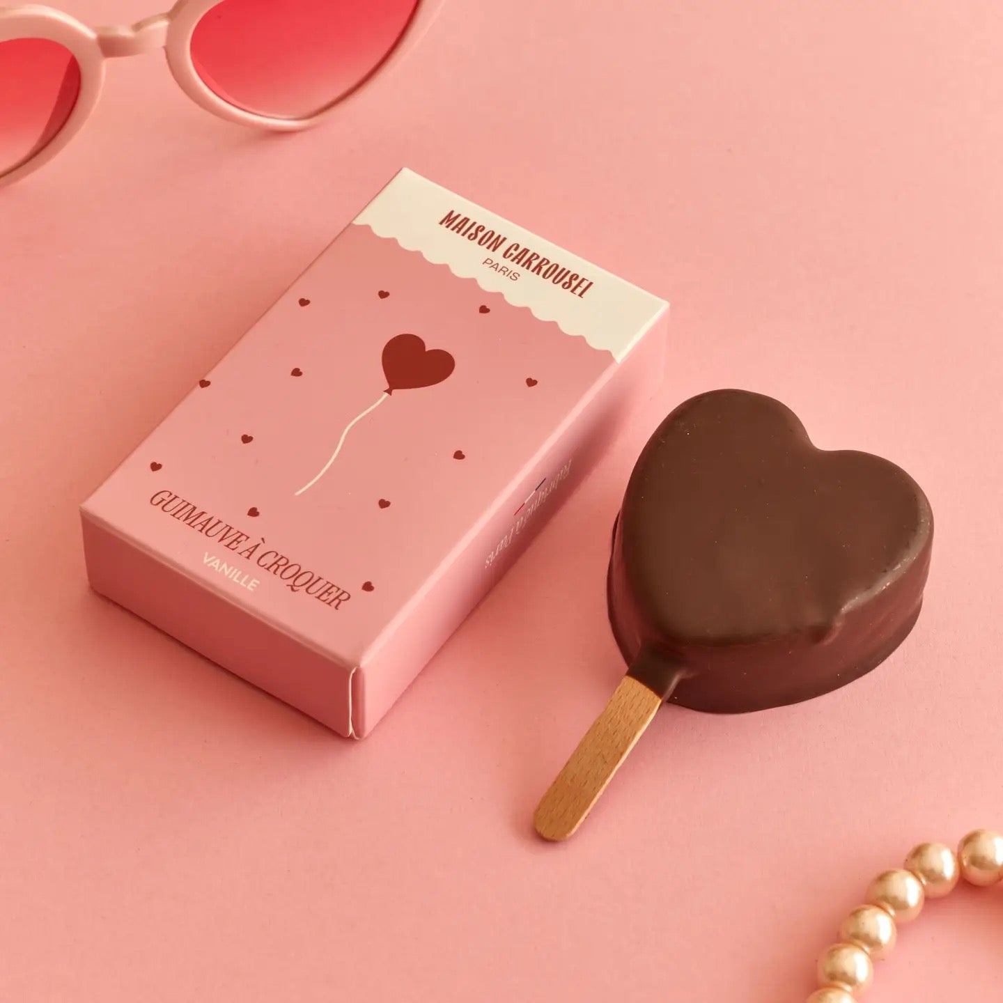 Herzförmiges Schokolade am Stiel neben einer rosa Verpackung mit der Aufschrift 'MAISON CARROUSEL PARIS' und 'GUIMAUVE À CROQUER VANILLE'.