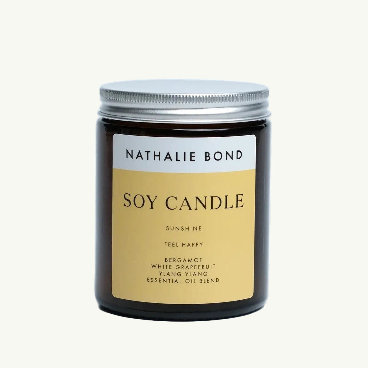 Braunes Glasglas mit silbernem Schraubdeckel, darauf ein gelb-beiger Aufkleber mit schwarzer Schrift 'NATHALIE BOND SOY CANDLE SUNSHINE FEEL HAPPY BERGAMOT WHITE GRAPEFRUIT YLANG YLANG ESSENTIAL OIL BLEND'. Duftkerze Sunshine