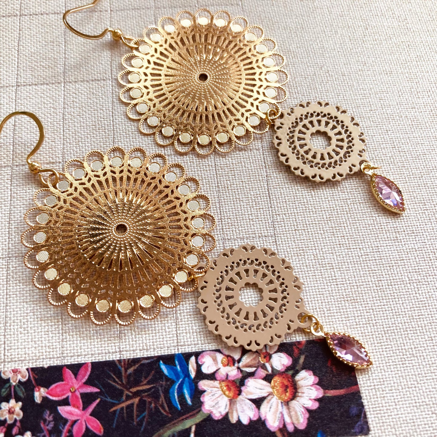 Goldfarbene Ohrringe mit filigranen, runden Mandala-Vebindern und tropfenförmigen, rosafarbenen Anhängern auf beigem Stoff