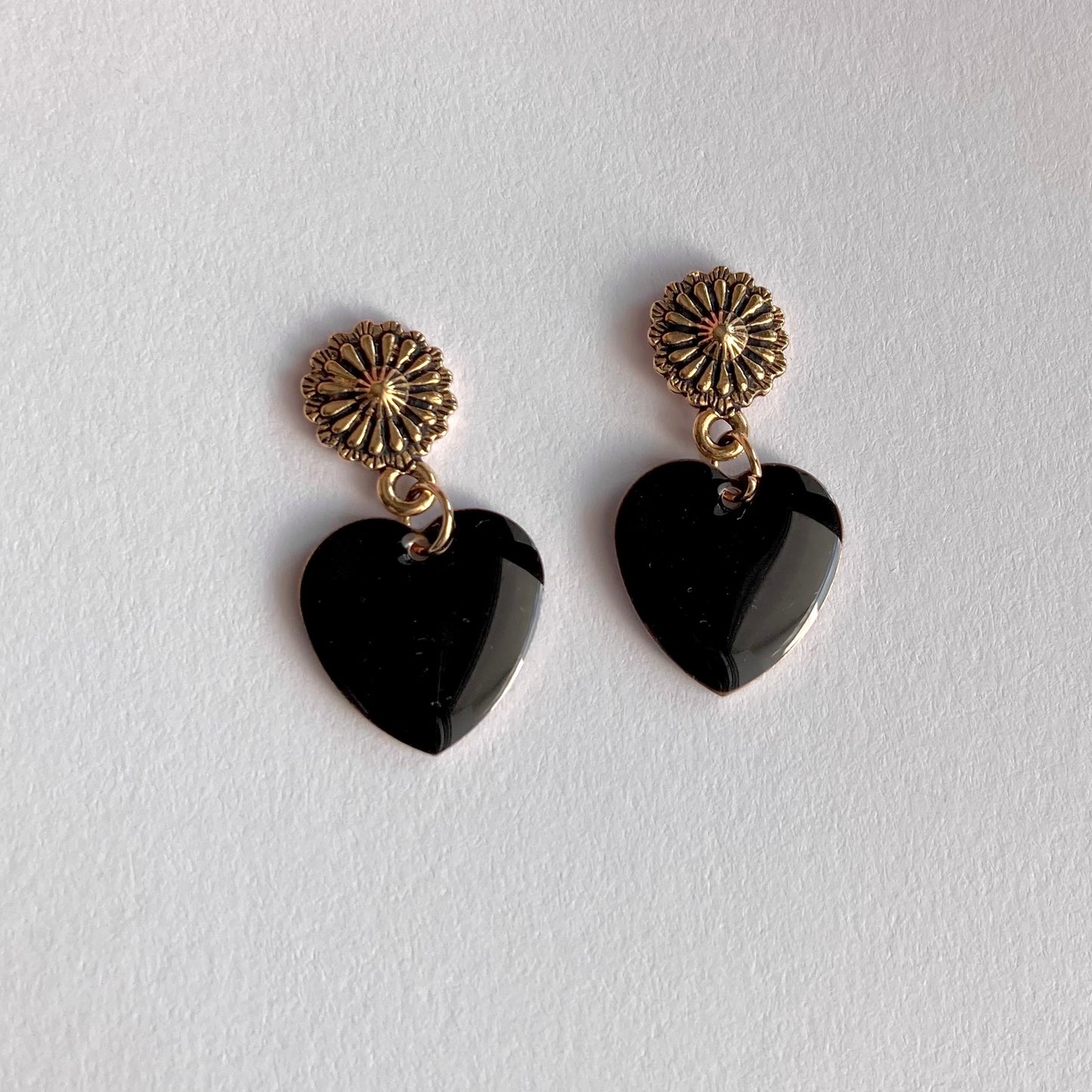 Ohrringe mit goldfarbenen, blumenförmigen Steckern und schwarzen, herzförmigen Anhängern auf weißem Untergrund