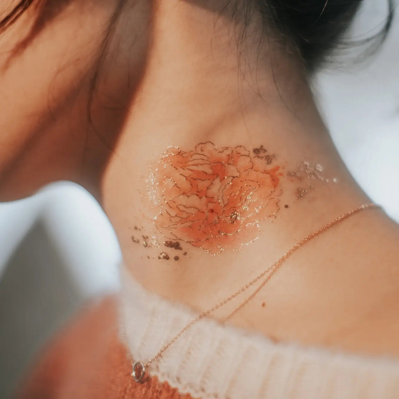 Blume als temporäres Tattoo auf dem Hals einer Frau mit heller Haut und braunen Haaren, die einen Pullover und eine dünne Halskette trägt