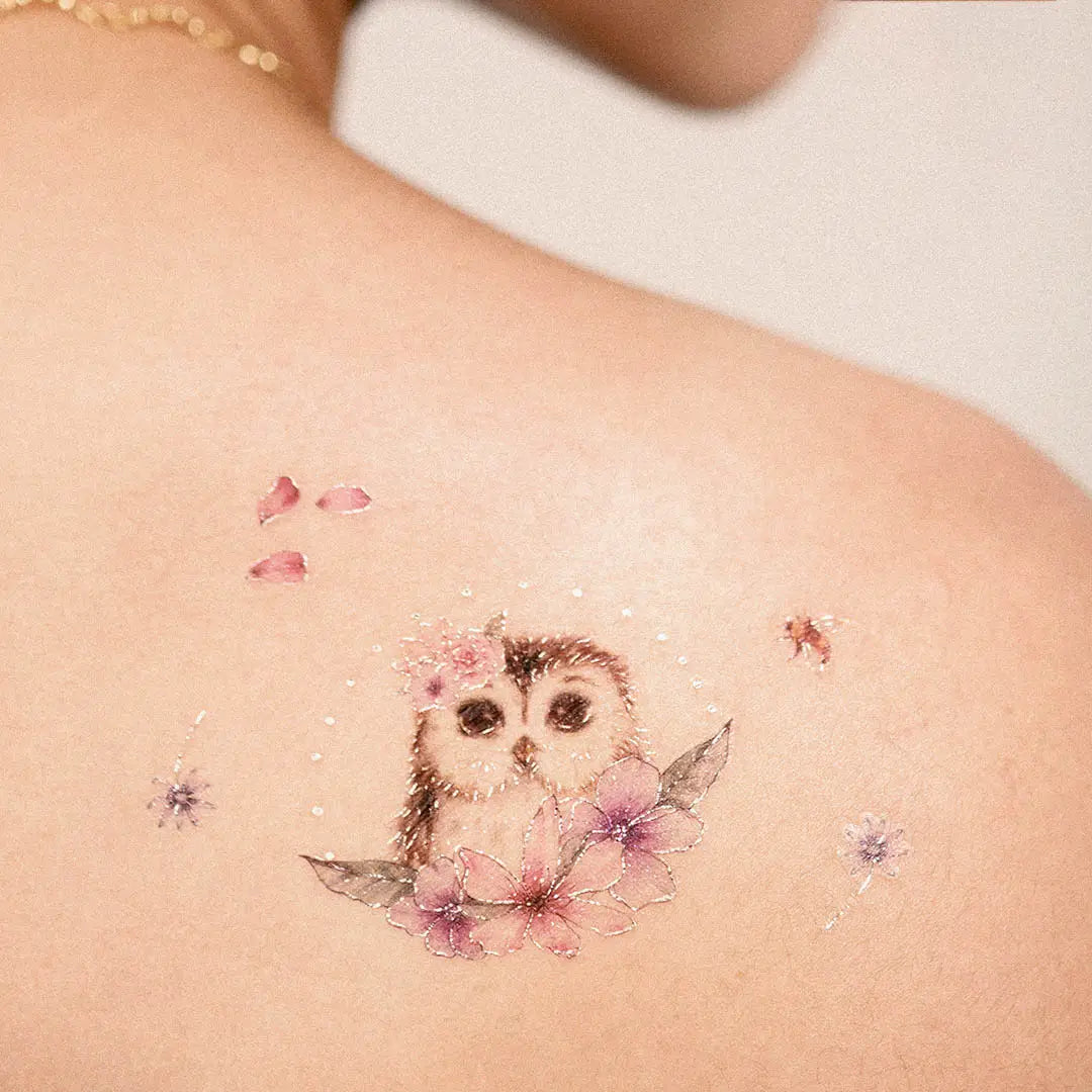 Tattoo auf Schulter mit zwei Eulen, umgeben von rosa Blüten und kleinen Blütenblättern