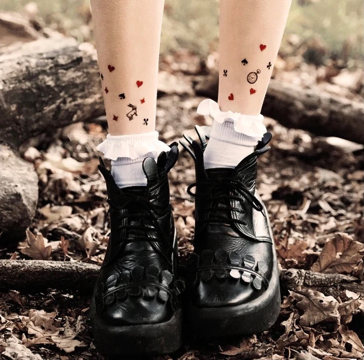 Beine mit weißen Socken und temporären Tattoos von Spielkartensymbolen, getragen mit schwarzen Plateau-Schnürschuhen auf Waldboden