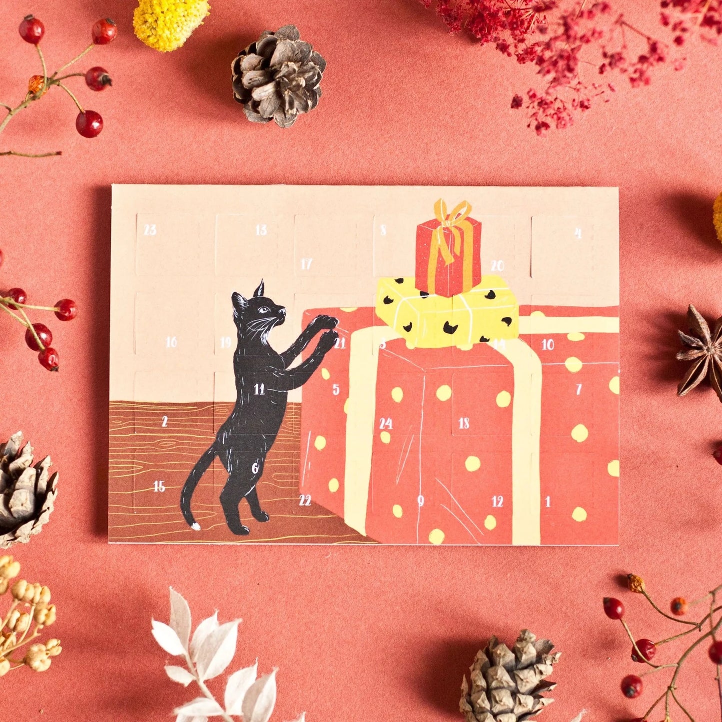 Adventskalender mit schwarzer Katze, die auf große rot-gelbe Geschenke mit Punkten springt, umgeben von Tannenzapfen und Beeren auf rotem Untergrund.