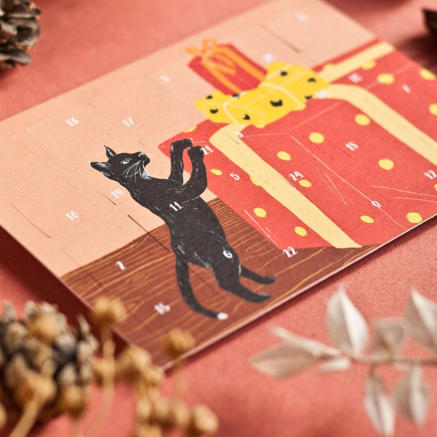 Adventskalender mit schwarzer Katze, die auf große rot-gelbe Geschenke mit Punkten springt, umgeben von Tannenzapfen und Beeren auf rotem Untergrund.