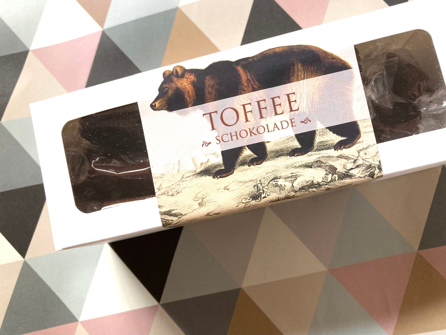 Weiße Schachtel mit durchsichtigen Fenstern, darin mehrere dunkle Toffee-Pralinen, Etikett mit braunem Bären und Schrift 'TOFFEE SCHOKOLADE'.