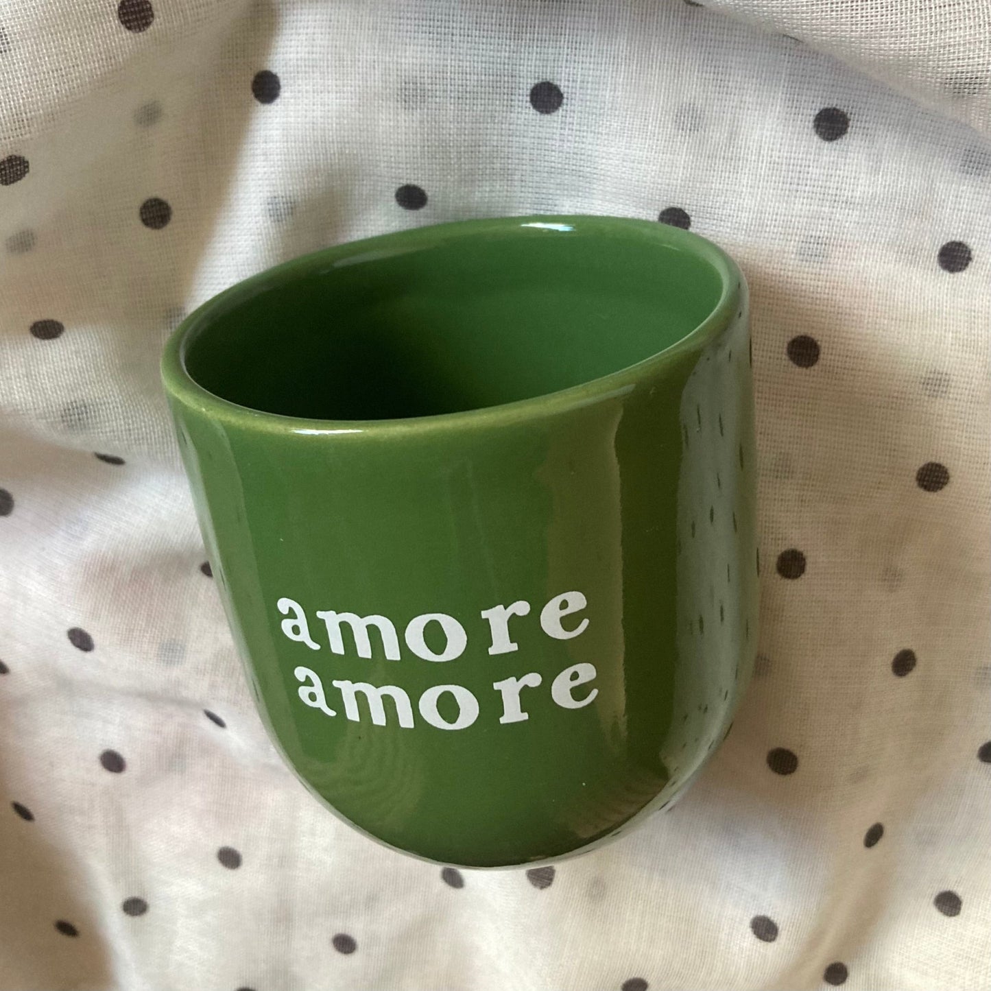 Ein glänzend grüner Keramikbecher mit der Aufschrift „Amore Amore“ in weißen Kleinbuchstaben, auf einem weißen Stoff mit schwarzem Tupfenmuster.