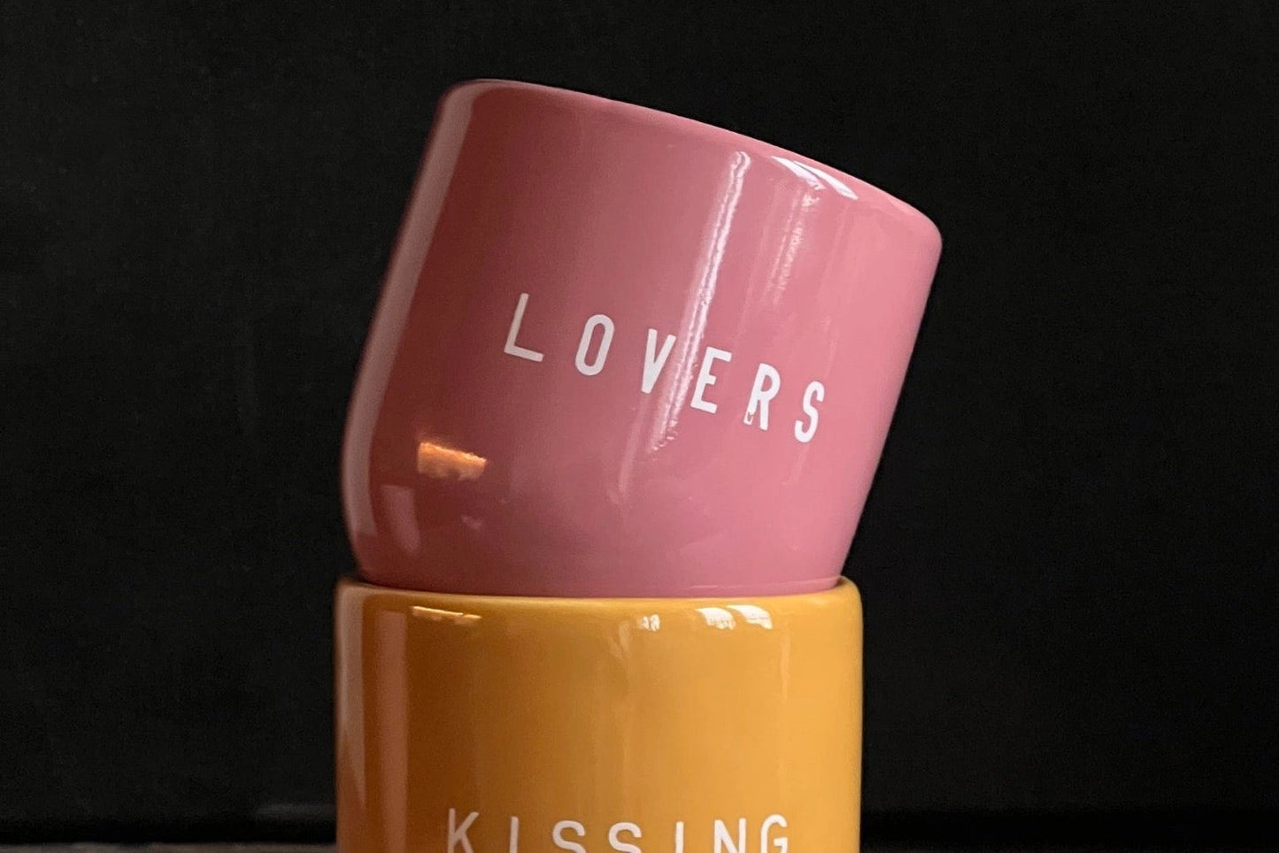 Ein rosa Keramikbecher mit weißer Aufschrift „LOVERS” auf schwarzem Hintergrund.