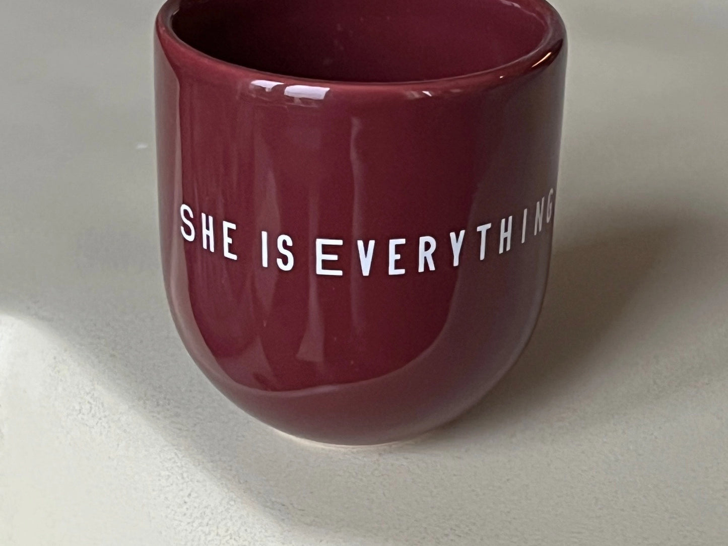 Ein weinroter Keramikbecher mit weißer Aufschrift „SHE IS EVERYTHING“ auf hellem Hintergrund.