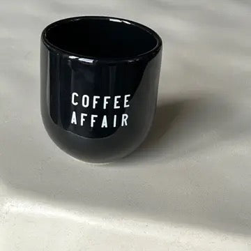 Ein schwarzer Keramikbecher mit weißer Aufschrift „COFFEE AFFAIR“ auf hellem Hintergrund.