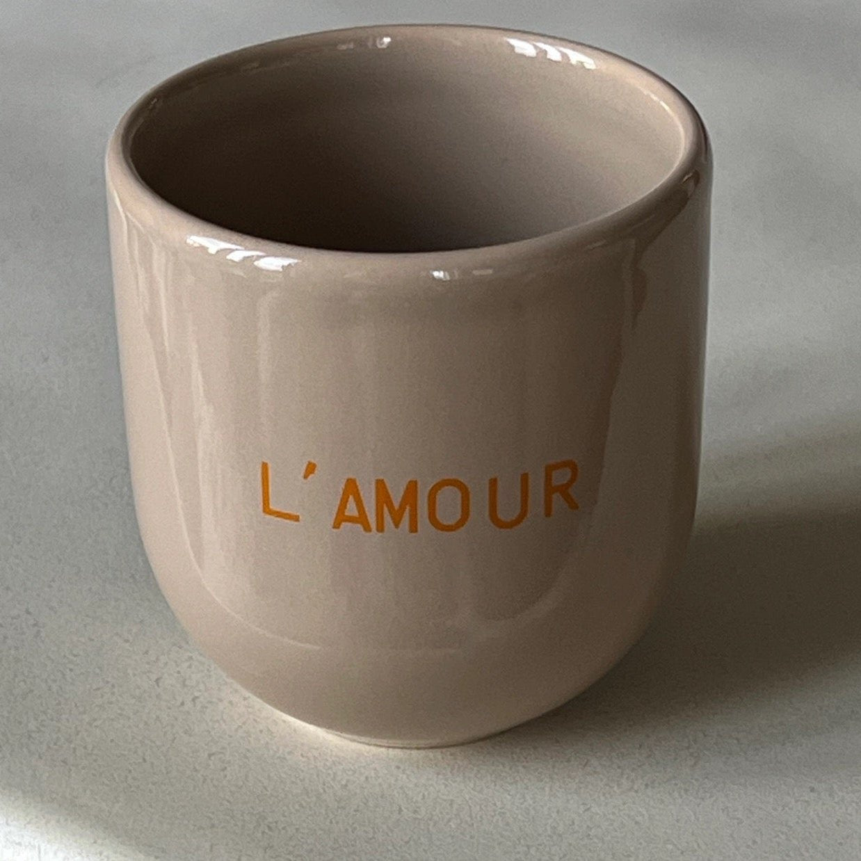 Ein hellbrauner Keramikbecher mit orangefarbener Aufschrift „L’AMOUR“ auf grauem Hintergrund