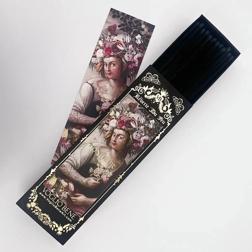 Schwarze Verpackung mit goldener Verzierung und Text 'Reverie Du Feu' enthält schwarze Streichhölzer, daneben eine Karte mit einem Porträt einer Frau mit Blumen im Haar