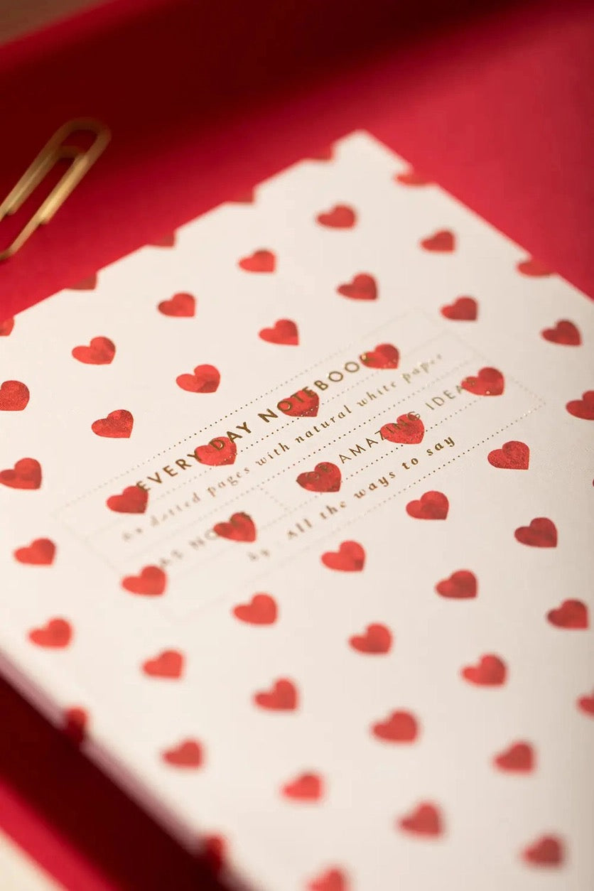 Nahaufnahme eines Notizhefts mit Herzmuster, das schräg auf einer roten Oberfläche liegt. Die Textur des Papiers und die glänzende Goldprägung des Titels sind im Fokus sichtbar, daneben liegt eine goldene Büroklammer.