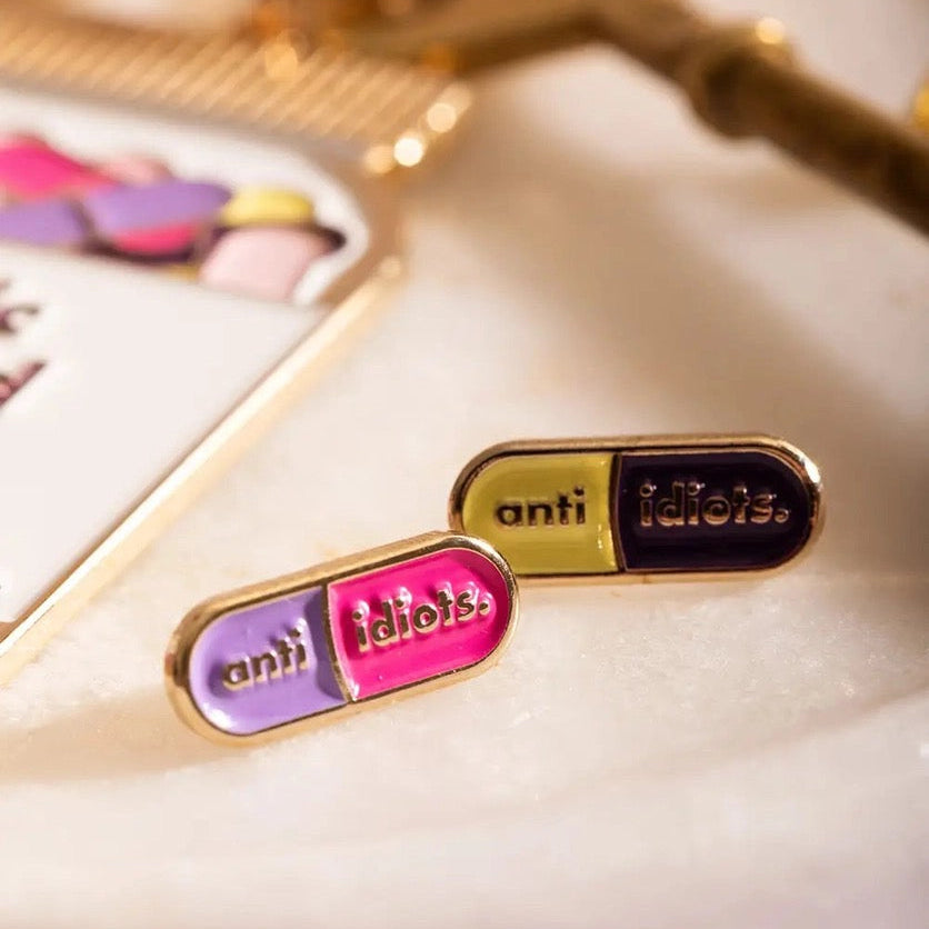 Zwei Emaille-Pins in Pillenform (lila/pink und gelb/dunkelblau) mit der Aufschrift „anti idiots“