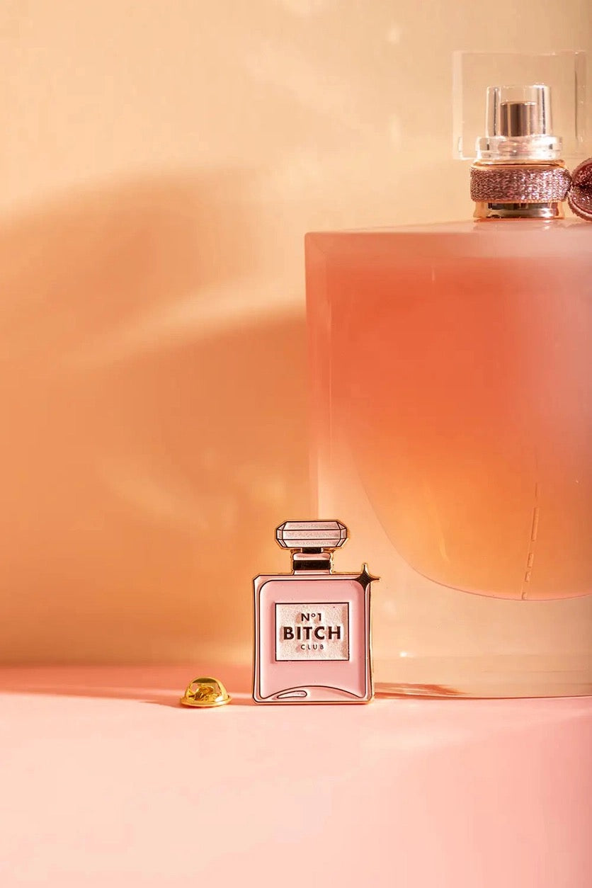 Duftet nach Selbstbewusstsein. Dieser Pin in Form eines luxuriösen Parfumflakons trägt das Label „N°1 Bitch Club“. Ein ironisches und zugleich glamouröses Statement-Piece für alle, die ihre Ecken und Kanten mit Stolz tragen.