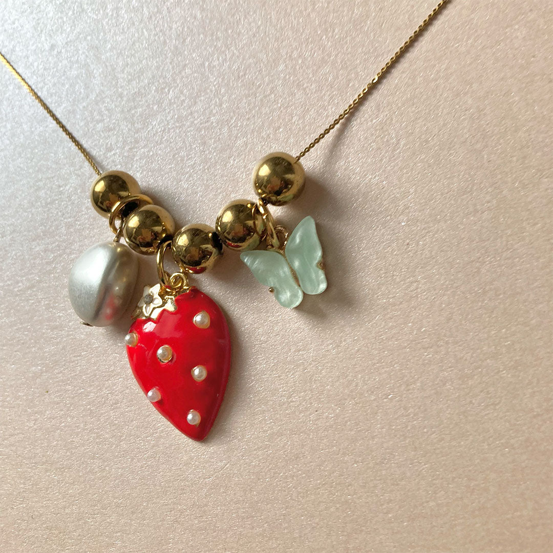 Halskette mit goldenen Kugeln, rotem Erdbeeranhänger mit weißen Punkten, weißem Perlenanhänger und hellgrünem Schmetterlingsanhänger.