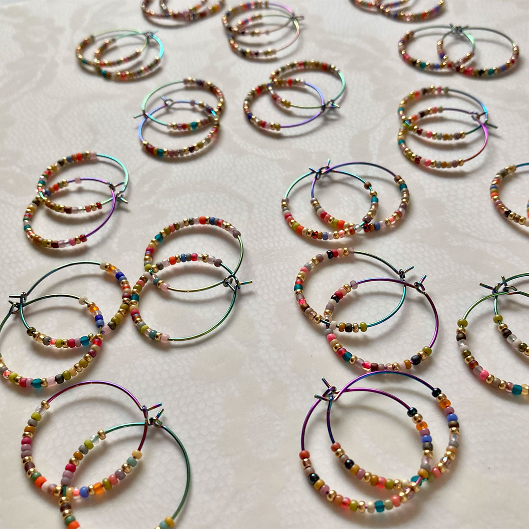 Bunte Perlen-Creolen in verschiedenen Farben auf einem hellen Untergrund. Die handgefertigten Ohrringe zeigen eine bunte Mischung aus Glasperlen und metallischen Akzenten an regenbogenfarbenen Metallreifen.