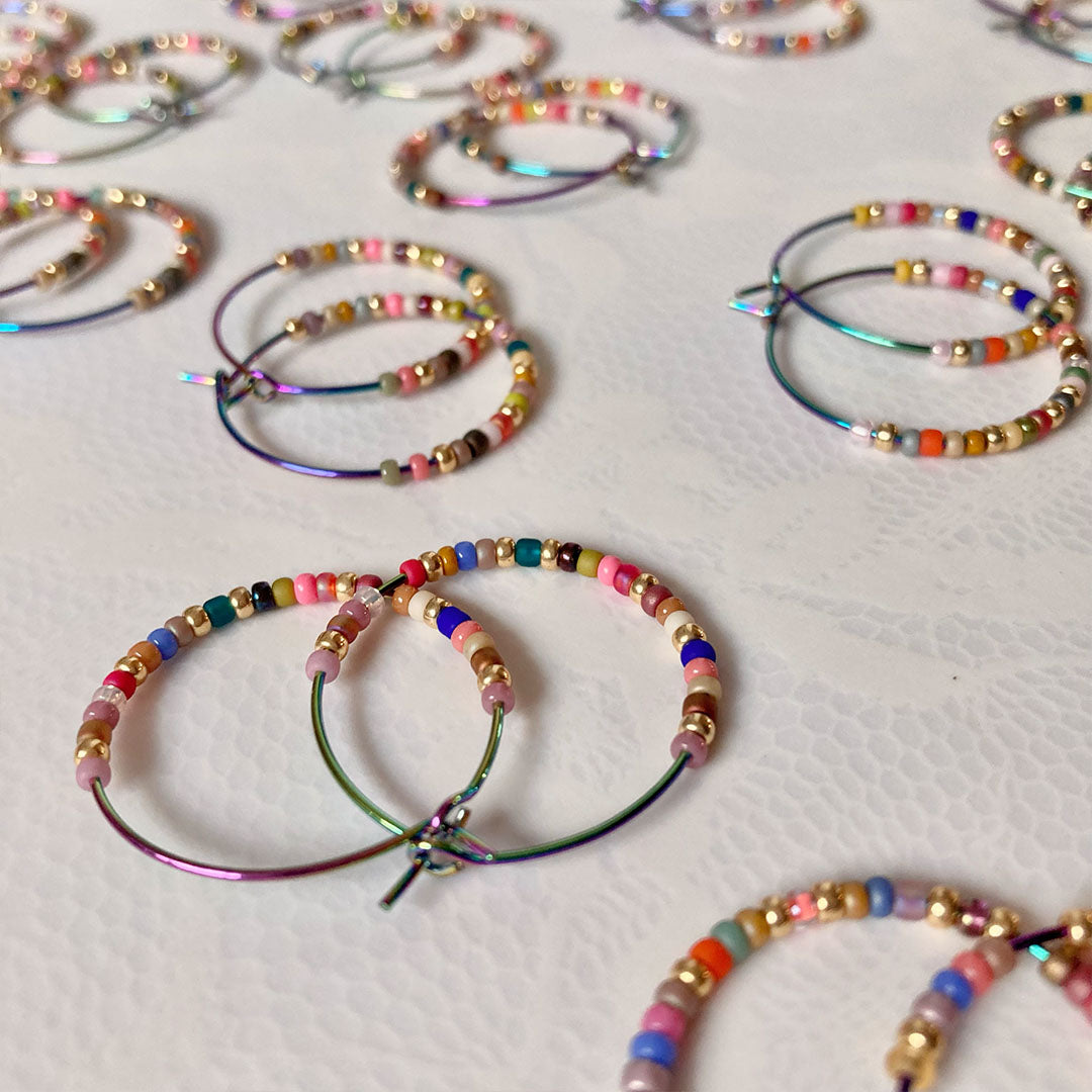 Bunte Perlen-Creolen in verschiedenen Farben auf einem hellen Untergrund. Die handgefertigten Ohrringe zeigen eine bunte Mischung aus Glasperlen und metallischen Akzenten an regenbogenfarbenen Metallreifen.