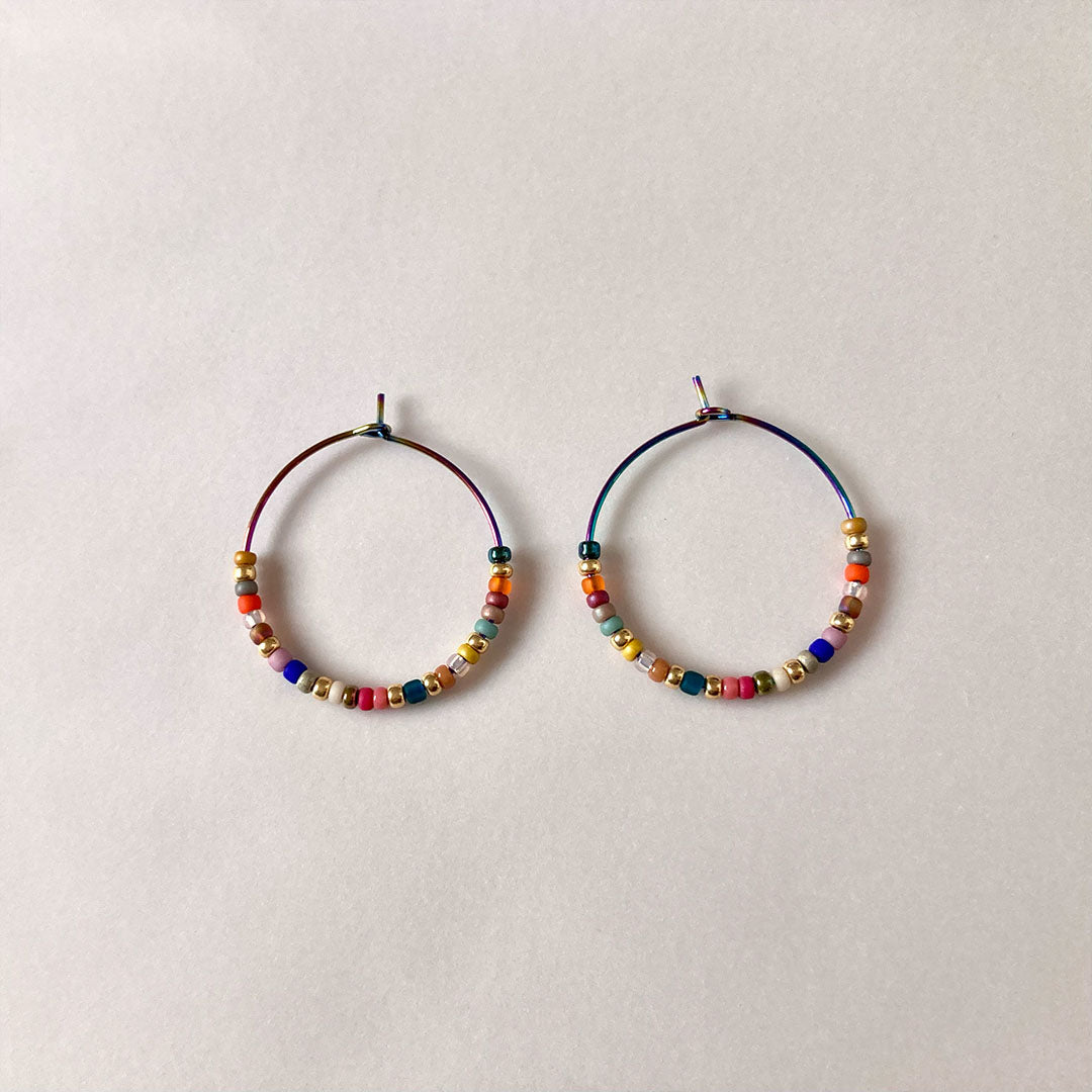 Bunte Perlen-Creolen in verschiedenen Farben auf einem hellen Untergrund. Die handgefertigten Ohrringe zeigen eine bunte Mischung aus Glasperlen und metallischen Akzenten an regenbogenfarbenen Metallreifen.