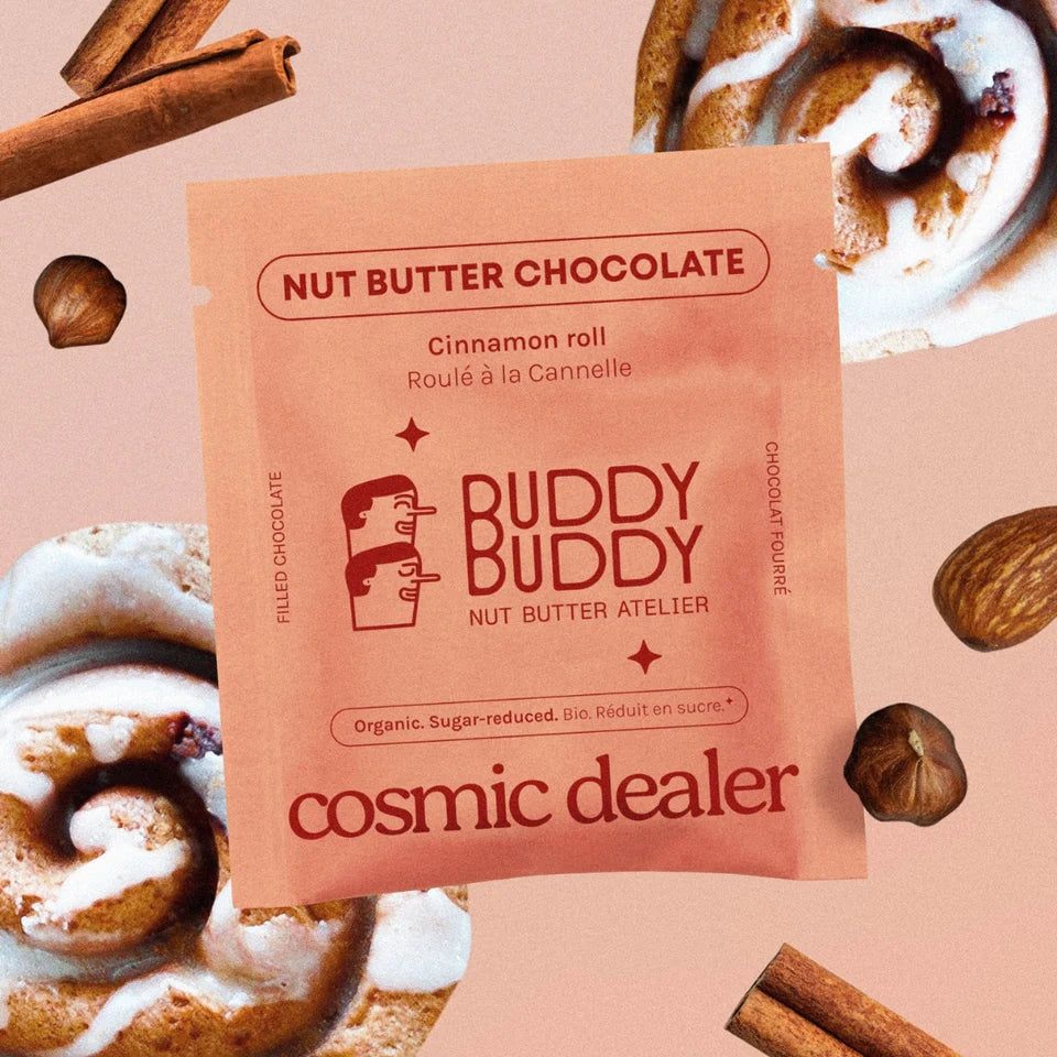 Cosmic Dealer x Buddy Buddy Nut Butter Chocolate Cinnamon Roll Verpackung auf lachsfarbenem Hintergrund, umgeben von Zimtstangen, Haselnüssen und Mandeln.
