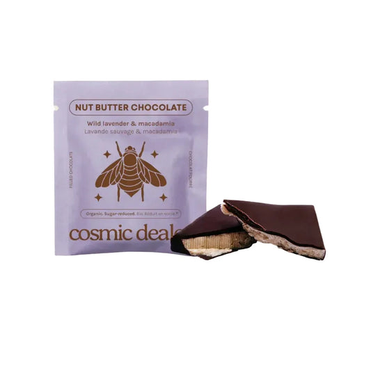 Eine durchgeschnittene dunkle Schokoladenpraline gibt den Blick frei auf die helle Macadamia-Nussbutter-Füllung, daneben die lila Cosmic Dealer Verpackung.