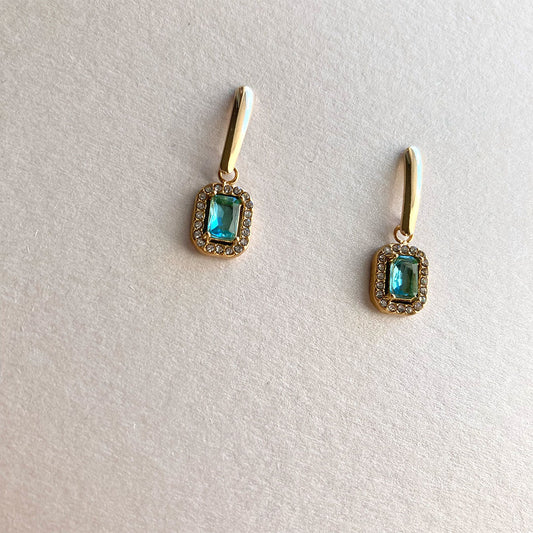 Minimalistische goldene Ohrhänger mit einem rechteckigen, hellblauen Schmuckstein. Der Stein ist von einem glitzernden Kranz aus kleinen Zirkonia-Steinen umgeben und hängt an einem schlichten, polierten Goldbügel.