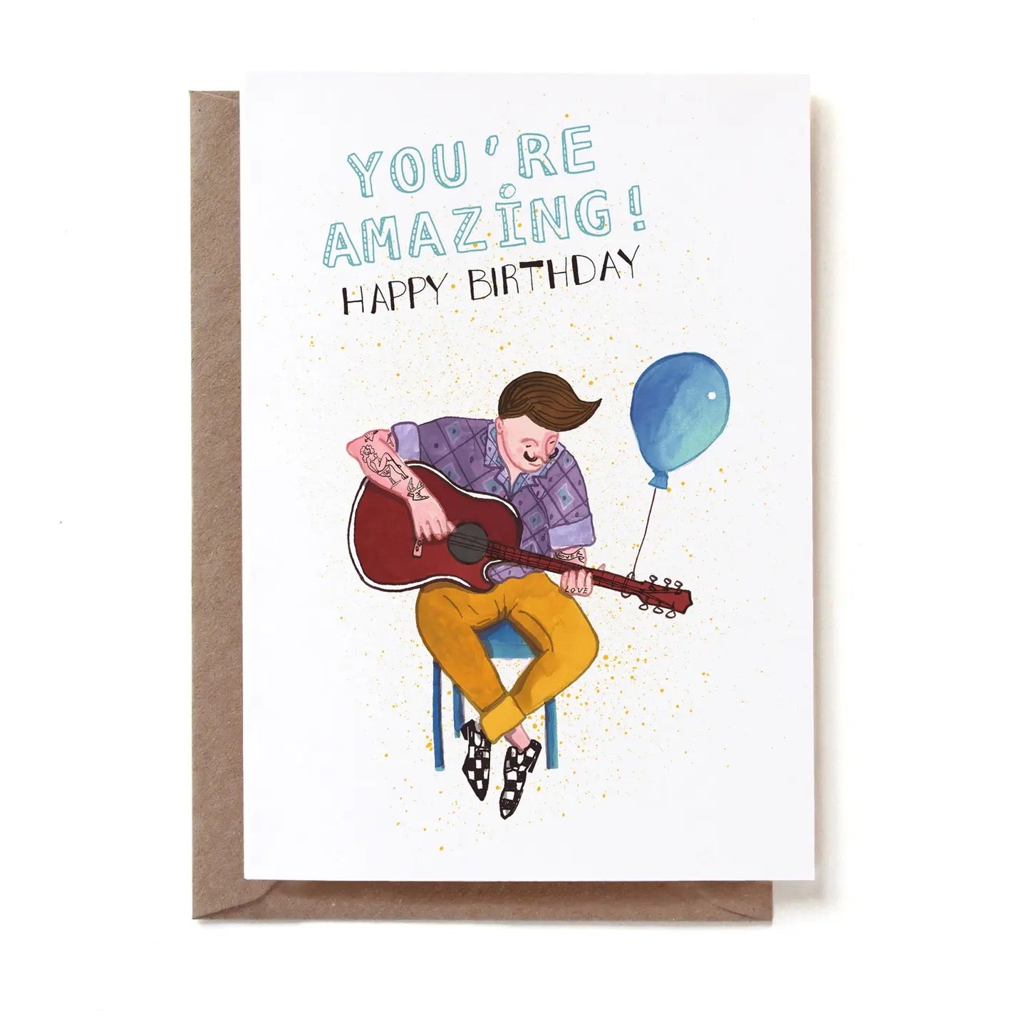 Eine illustrierte Geburtstagskarte mit der Aufschrift „YOU'RE AMAZING! HAPPY BIRTHDAY“. Ein Mann mit markanter Tolle und tätowierten Armen sitzt auf einem Hocker und spielt eine akustische Gitarre. Er trägt ein lila gemustertes Hemd, eine gelbe Hose und gemusterte Slipper. An den Hals der Gitarre ist ein schwebender blauer Luftballon gebunden. Der Hintergrund ist weiß mit feinen gelben Farbspritzern. Die Karte steckt in einem braunen Kraftpapier-Umschlag.