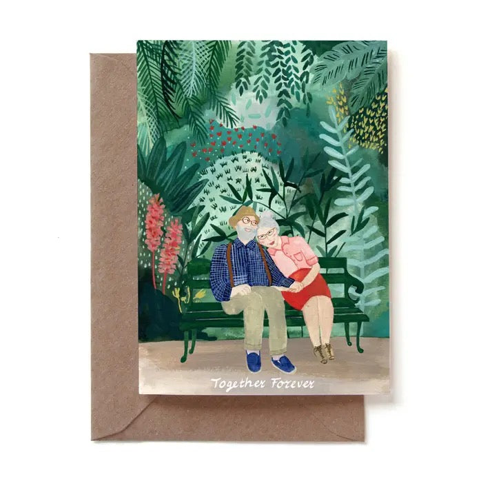 Eine illustrierte Grußkarte mit einem älteren Paar, das glücklich Hand in Hand auf einer grünen Parkbank sitzt. Der Mann trägt ein blau kariertes Hemd mit Hosenträgern, die Frau ein rosa Oberteil und einen roten Rock. Sie sind von einem dichten, grünen Garten mit Farnen und roten Blumen umgeben. Am unteren Rand steht der Text „Together Forever“. Die Karte steckt in einem braunen Kraftpapier-Umschlag.