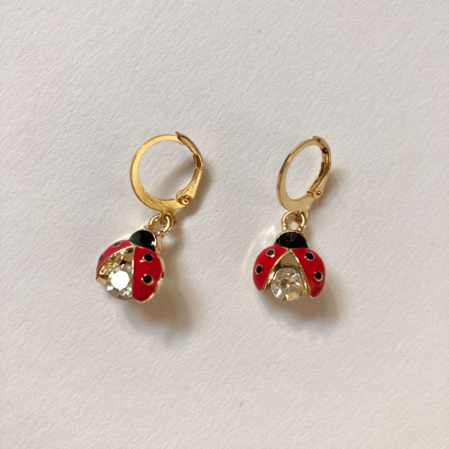 Goldene Creolen-Ohrringe mit Marienkäfer-Anhängern. Die Marienkäfer haben rote Flügel mit schwarzen Punkten und einen großen, funkelnden Kristall-Körper.