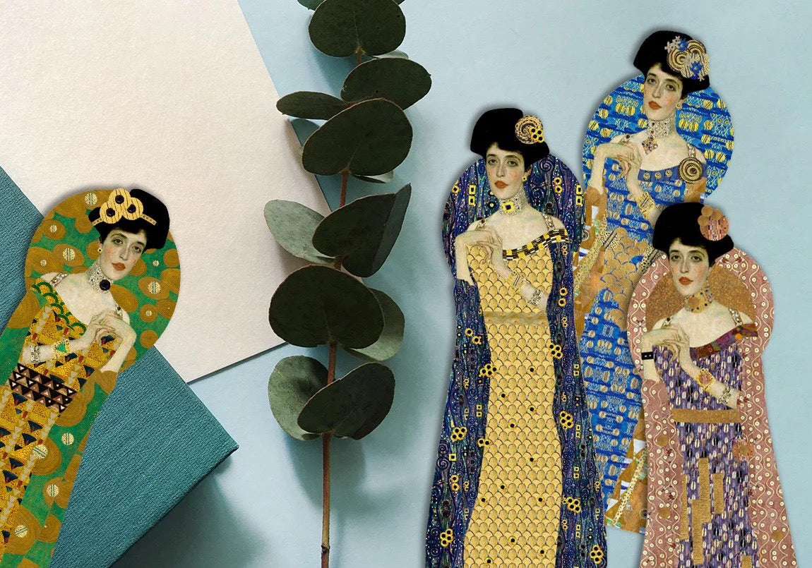 Flachbild-Arrangement von vier figürlich gestanzten Gustav-Klimt-Lesezeichen auf einem hellblauen Hintergrund. Neben den Lesezeichen liegt ein grüner Eukalyptuszweig, ein weißes Papierblatt und ein petrolfarbenes Buch. Die Lesezeichen zeigen das Porträt einer eleganten Frau im Jugendstil, gekleidet in gemusterten Roben in Grün, Gelb-Blau, leuchtendem Blau und Altrosa.