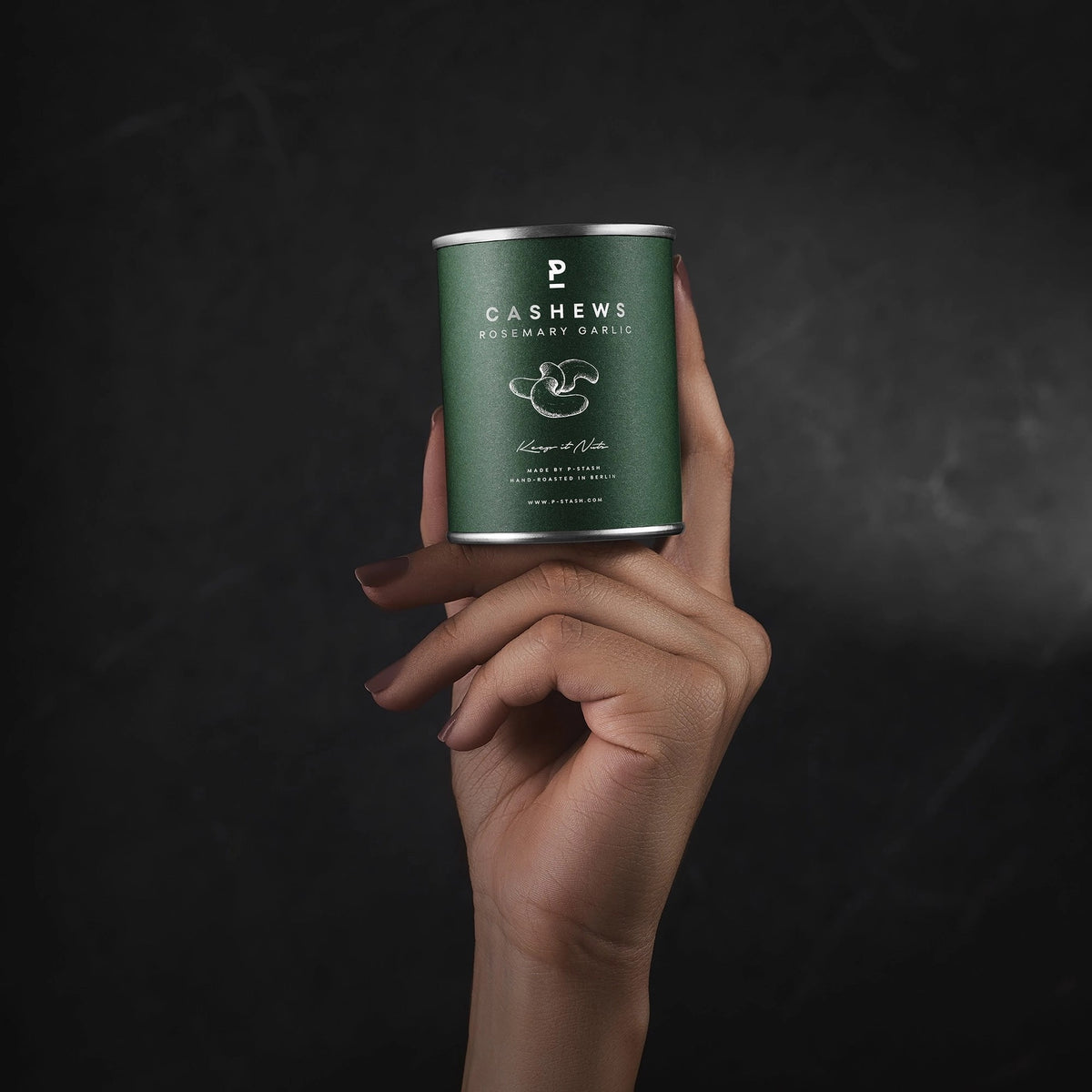 Eine Hand präsentiert eine dunkelgrüne P-Stash Dose für Rosemary-Garlic-Cashews aufrecht vor einem schlichten, dunkelgrauen Hintergrund.