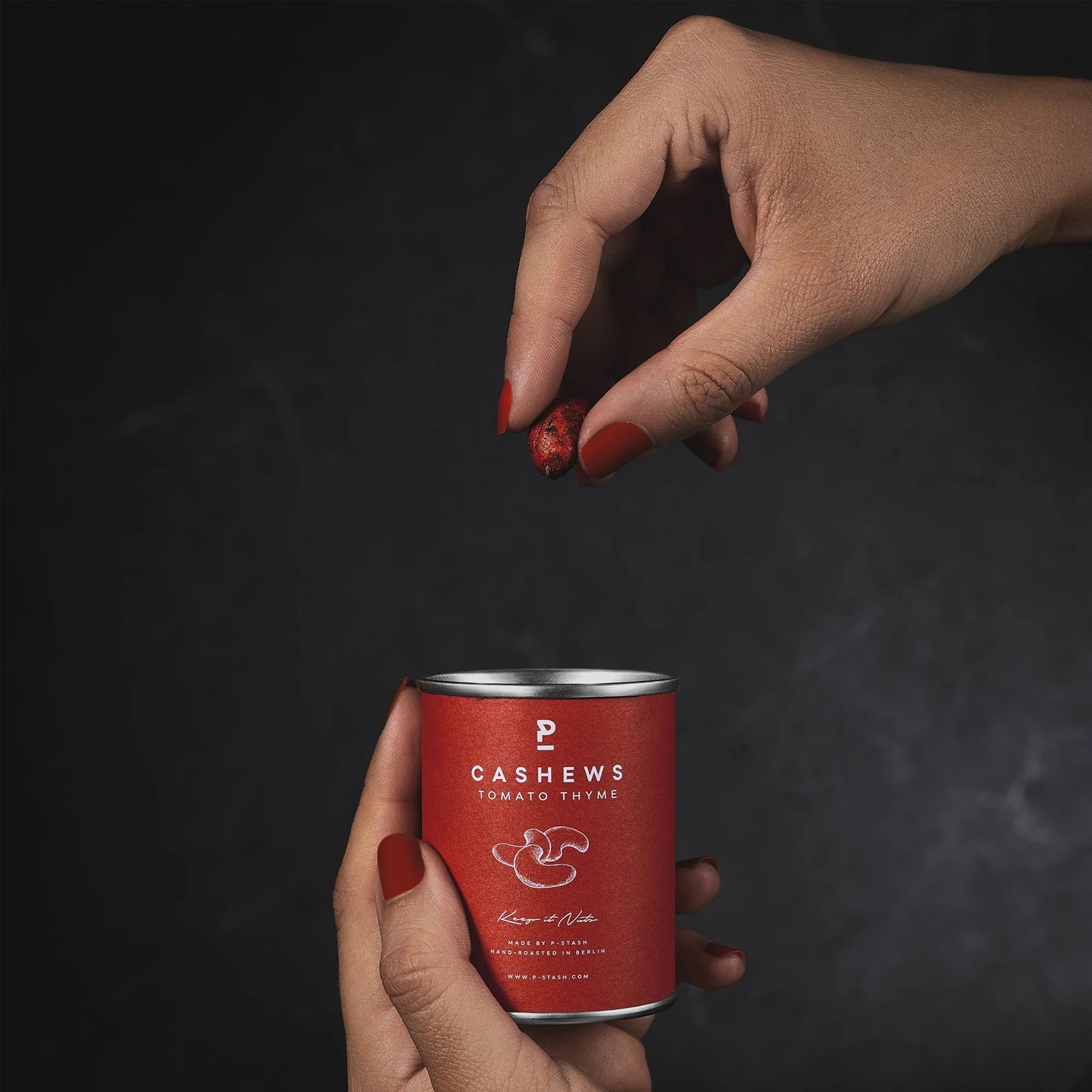 Hand mit roten Nägeln hält eine Dose Cashews mit Tomate-Thymian-Geschmack, andere Hand hält eine Cashewnuss