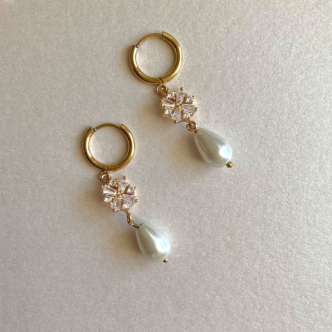 Ein Paar elegante, goldfarbene Creolen-Ohrringe auf weißem Grund. Die Ohrringe verfügen über einen abnehmbaren Anhänger, bestehend aus einem funkelnden, blumenförmigen Element aus Zirkonia-Steinen und einer darunter hängenden, tropfenförmigen weißen Perle.