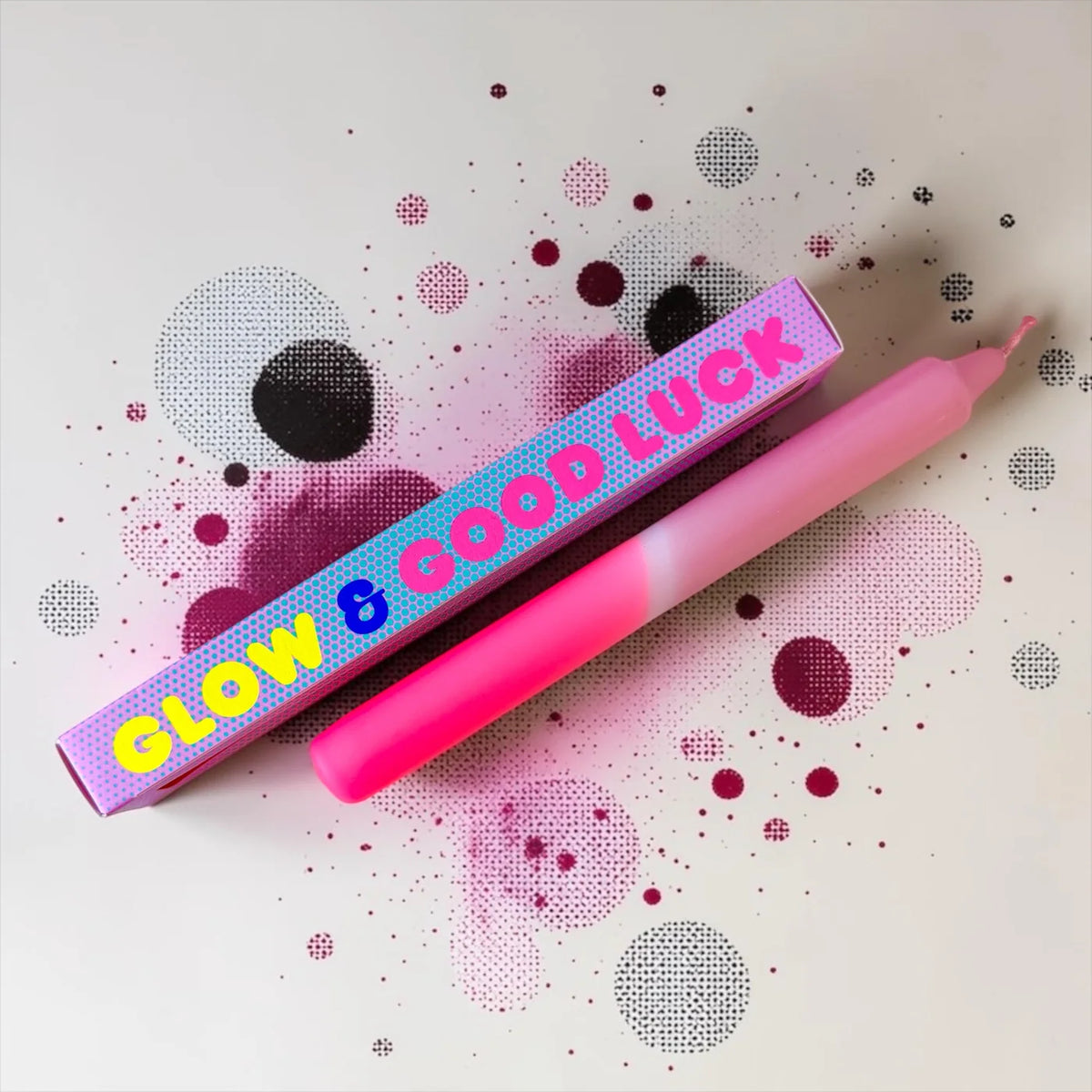 Eine pinke Dip-Dye-Stabkerze von Pink Stories mit sanftem Farbverlauf von hellem Neon-Rosa zu Altrosa, daneben liegt die passende bedruckte Geschenkbox mit der Aufschrift „GLOW & GOOD LUCK“.