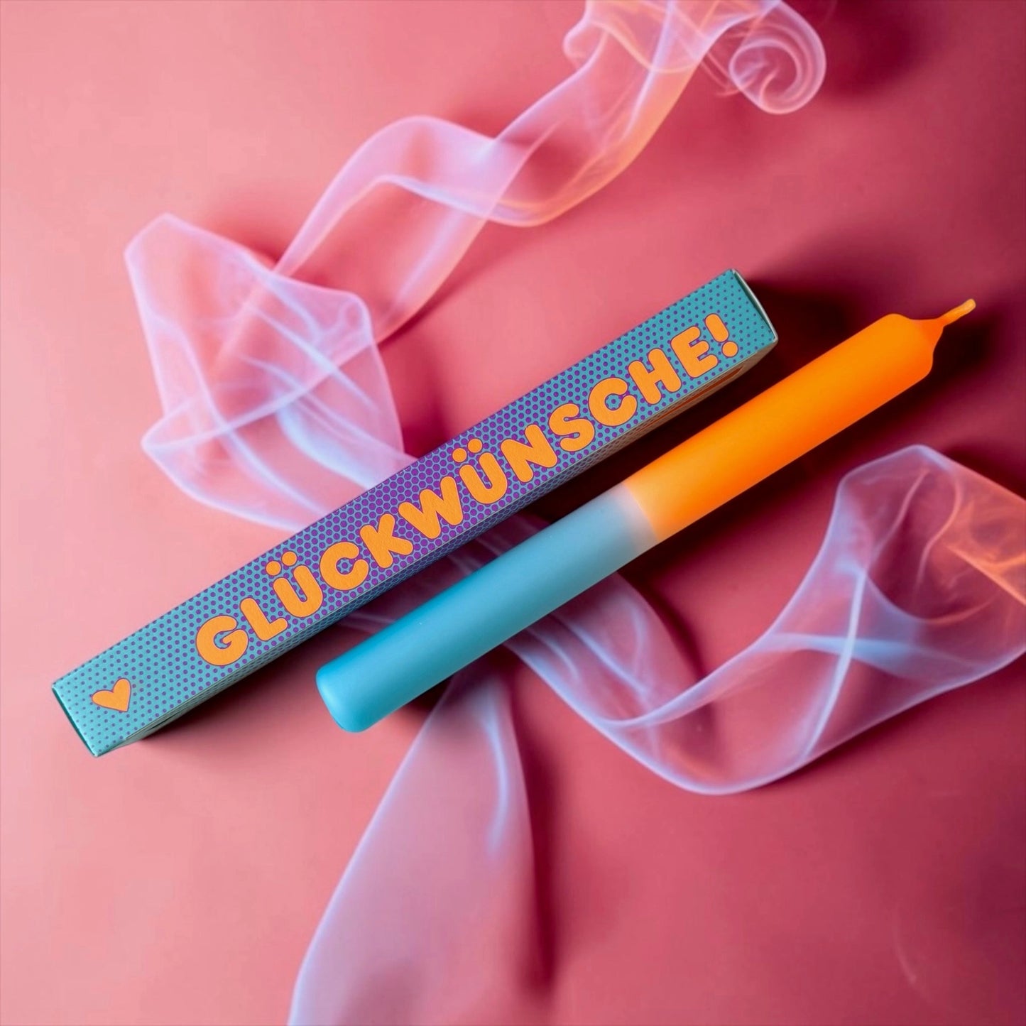 Zweifarbige Stabkerze in Türkis und Orange auf rosa Hintergrund mit durchscheinendem Schleierstoff. Daneben die türkisblau gepunktete Schachtel mit der orangen Aufschrift „GLÜCKWÜNSCHE!“.
