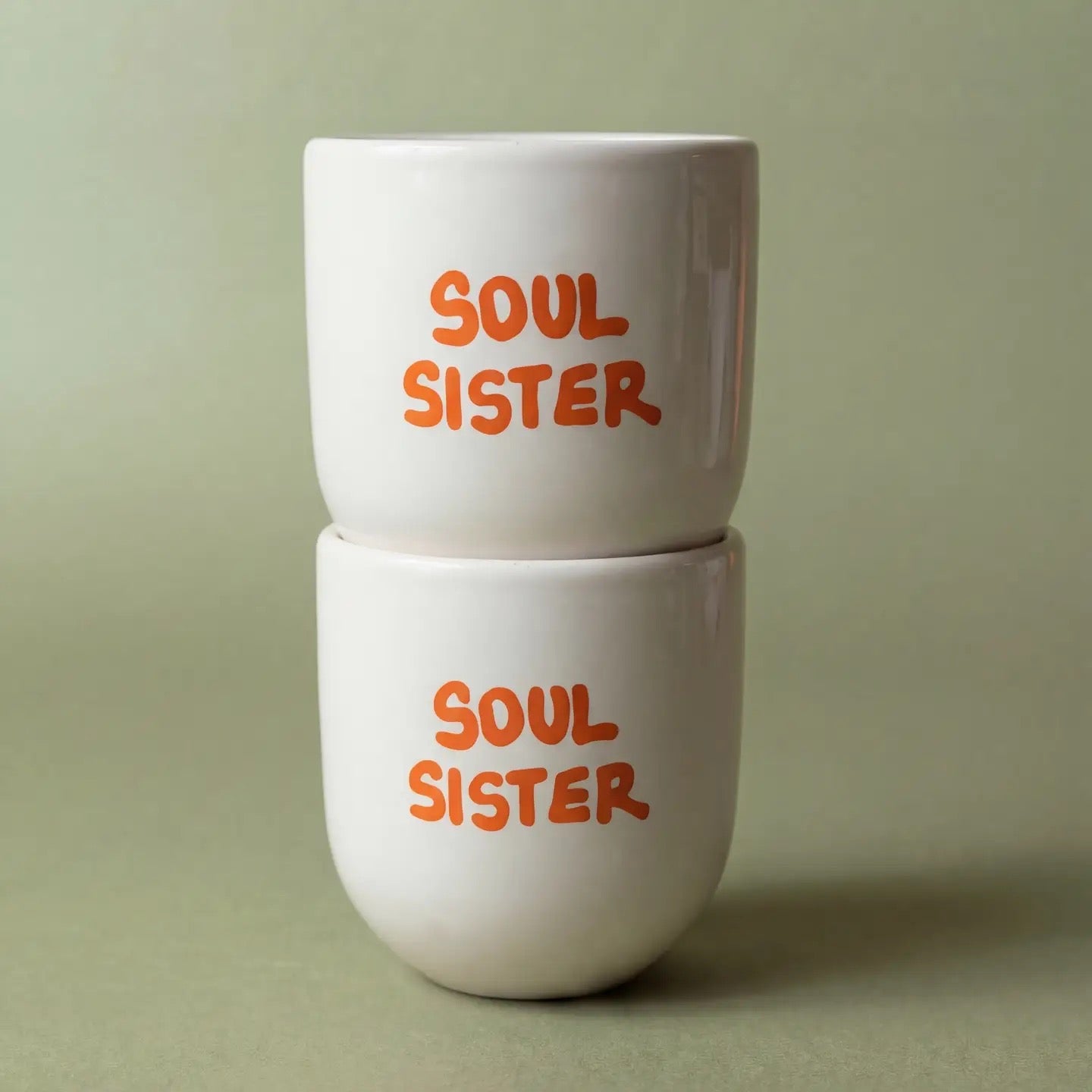 Zwei cremeweiße, gestapelte Keramikbecher von House of Style mit der orangefarbenen Aufschrift "Soul Sister" vor einem salbeigrünen Hintergrund.