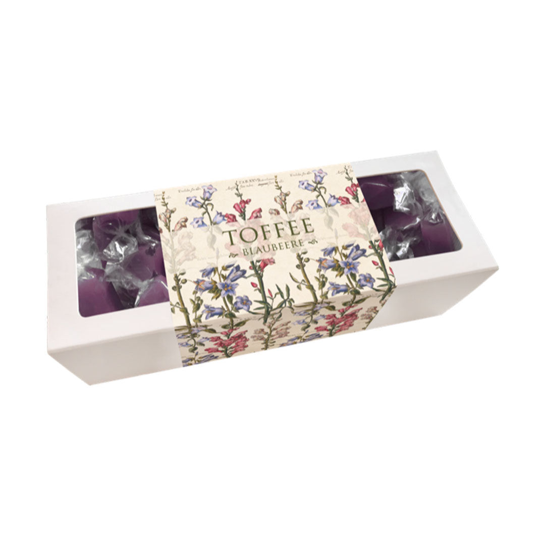 Produktfoto einer weißen Pralinenschachtel mit Blaubeer-Toffees. Die dekorative Banderole im Vintage-Stil ist mit blauen Glockenblumen und botanischen Skizzen verziert. Durch das Fenster sind die violetten, einzeln verpackten Toffees sichtbar. Schriftzug: „TOFFEE BLAUBEERE“.