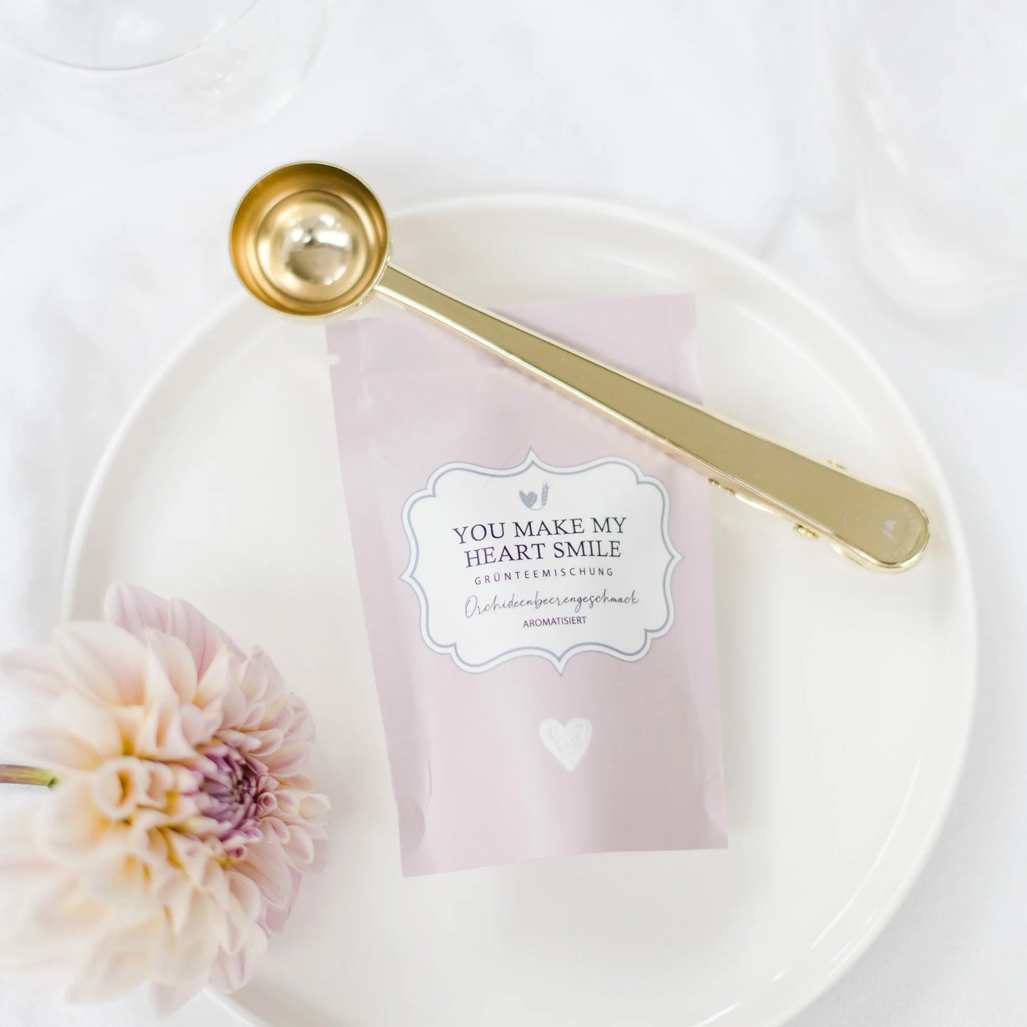 Pinke Teeverpackung „YOU MAKE MY HEART SMILE“ auf einem weißen Teller, dekoriert mit einer passenden Blüte und einem goldenen Dosierlöffel